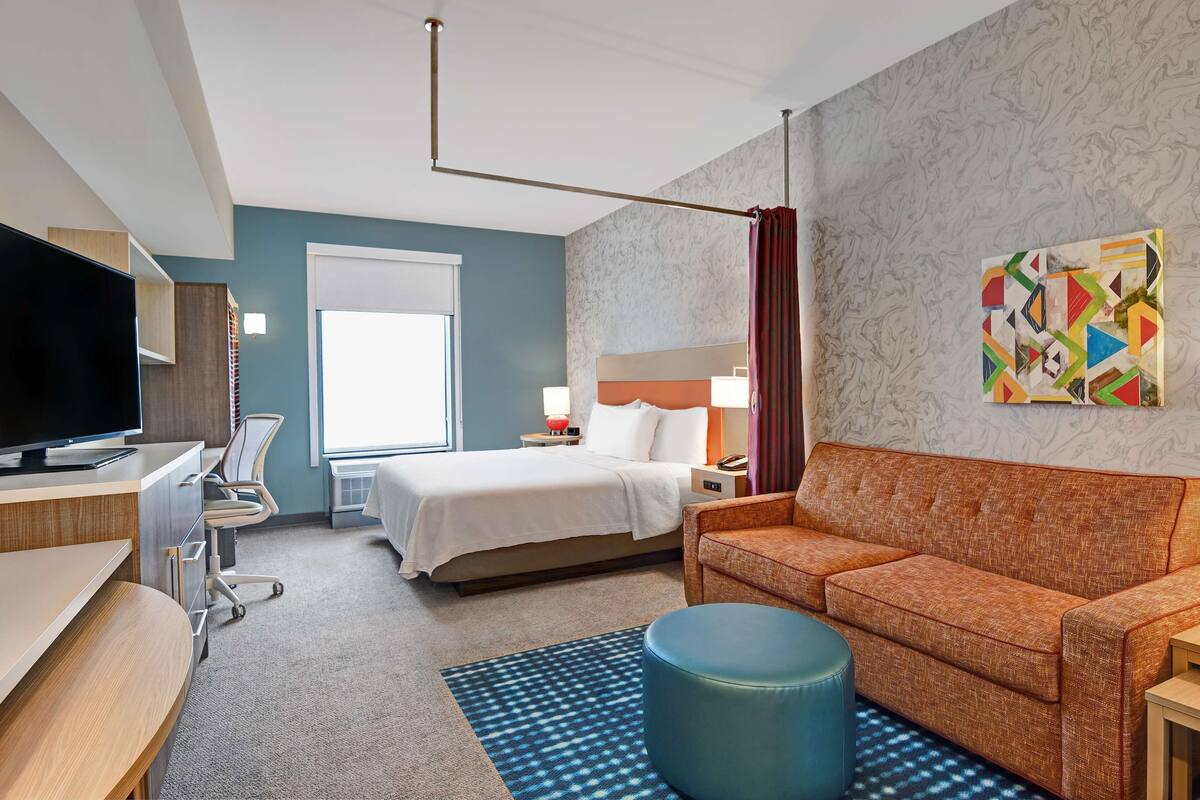 Ein Hotelzimmer mit einem Bett, einem Schreibtisch mit Fernseher, einer Couch, einem Sessel, einer Lampe und einem Bild an der Wand.
