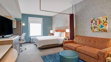 A hotel room with a bed, a desk with a TV, a sofa, a chair, a lamp, and a painting on the wall.