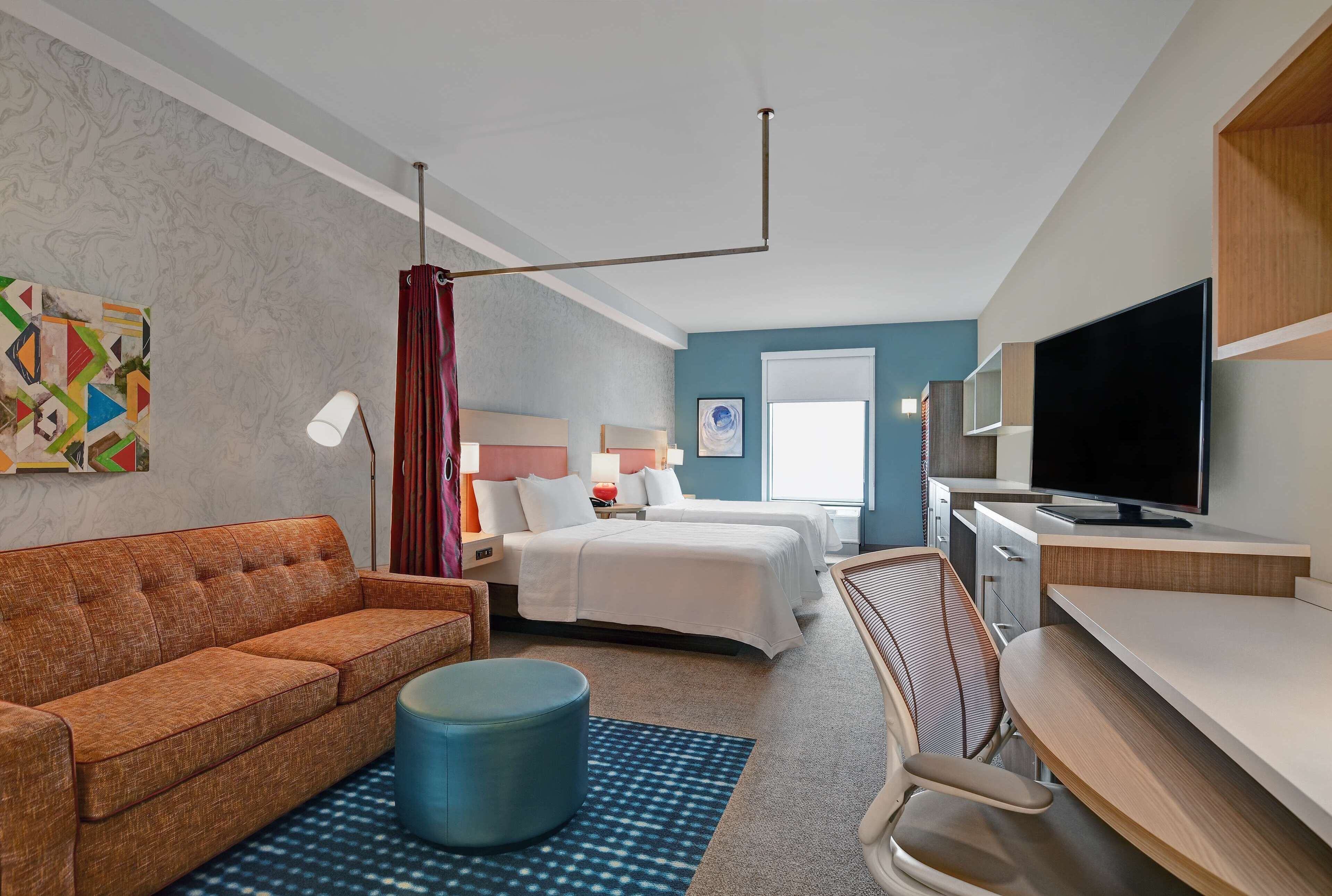 A hotel room with a sofa, two beds, a desk, a TV, and a painting on the wall.