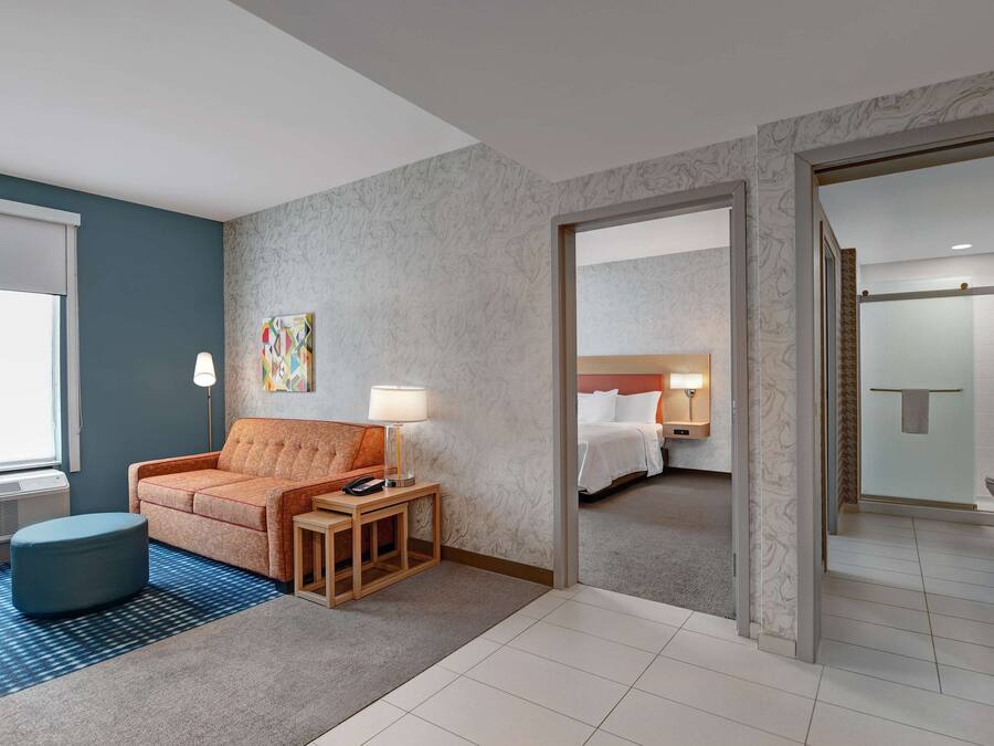 A hotel room with a sofa, a small table with a lamp, a blue ottoman, a bed with white bedding, and a bathroom visible through an open door.