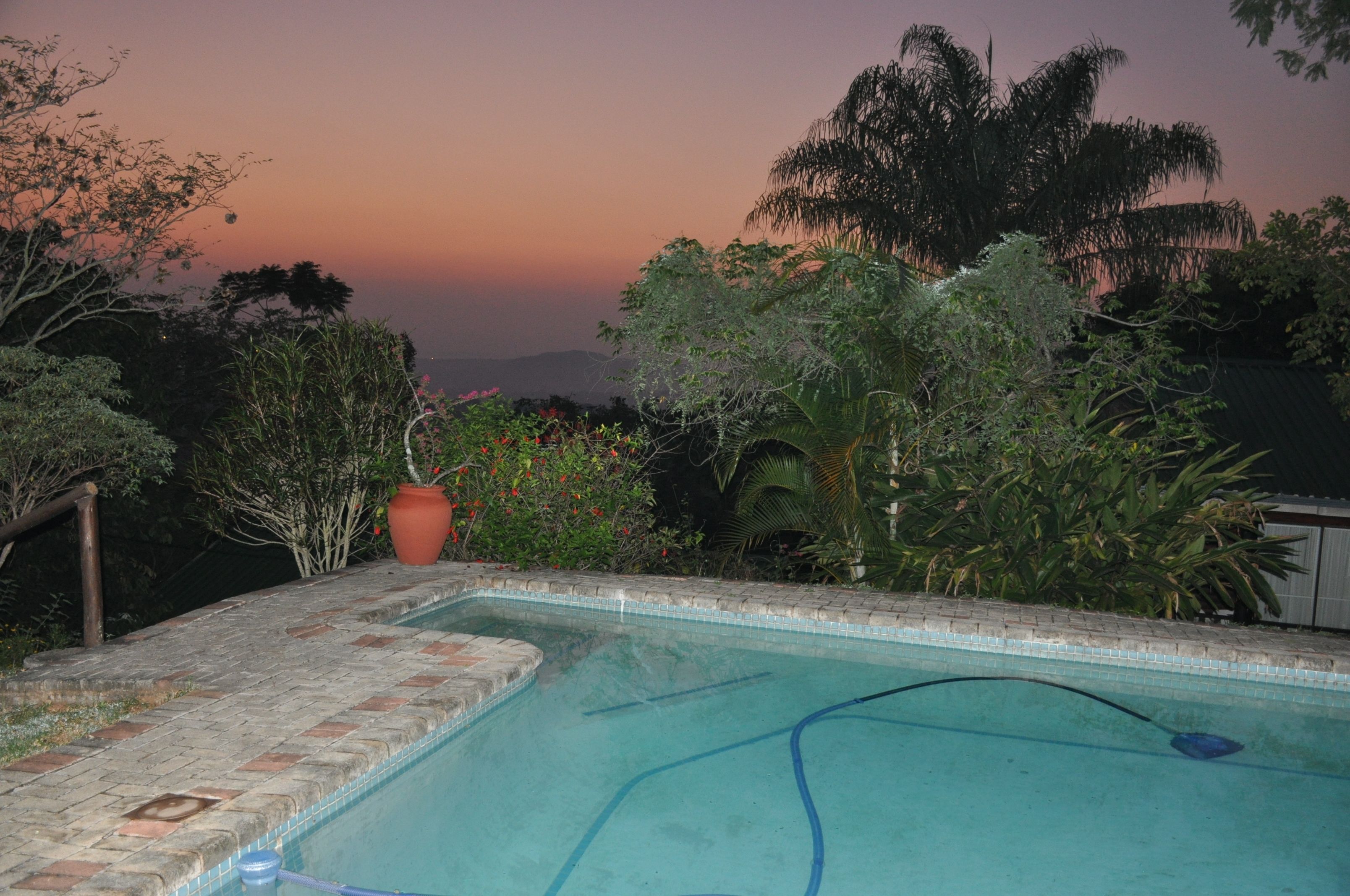 Una piscina con borde de ladrillo, rodeada de exuberante vegetación y vistas a colinas distantes al atardecer.
