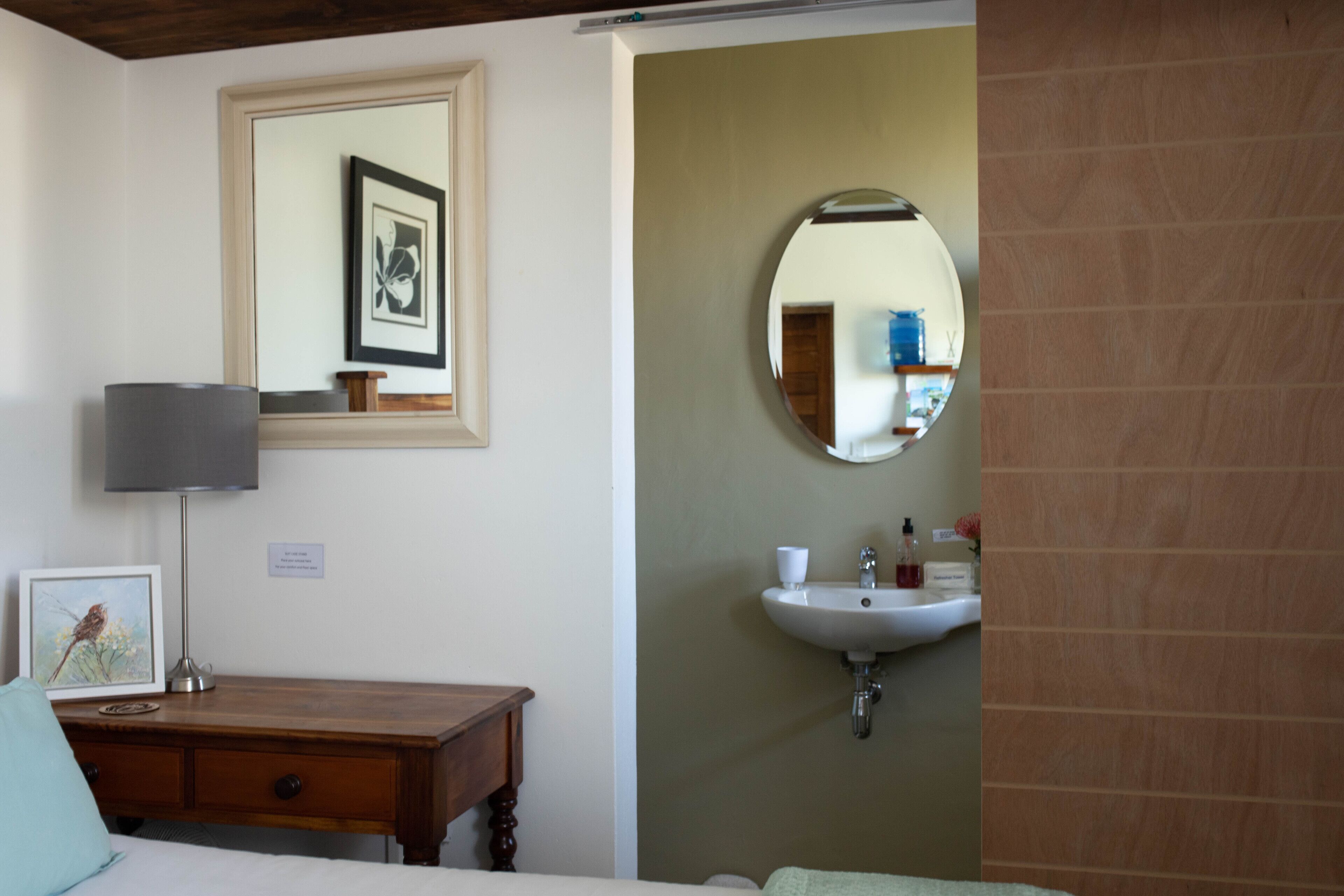A bedroom with a wooden dresser, a bedside lamp, a framed picture, a round mirror, and a white sink.
