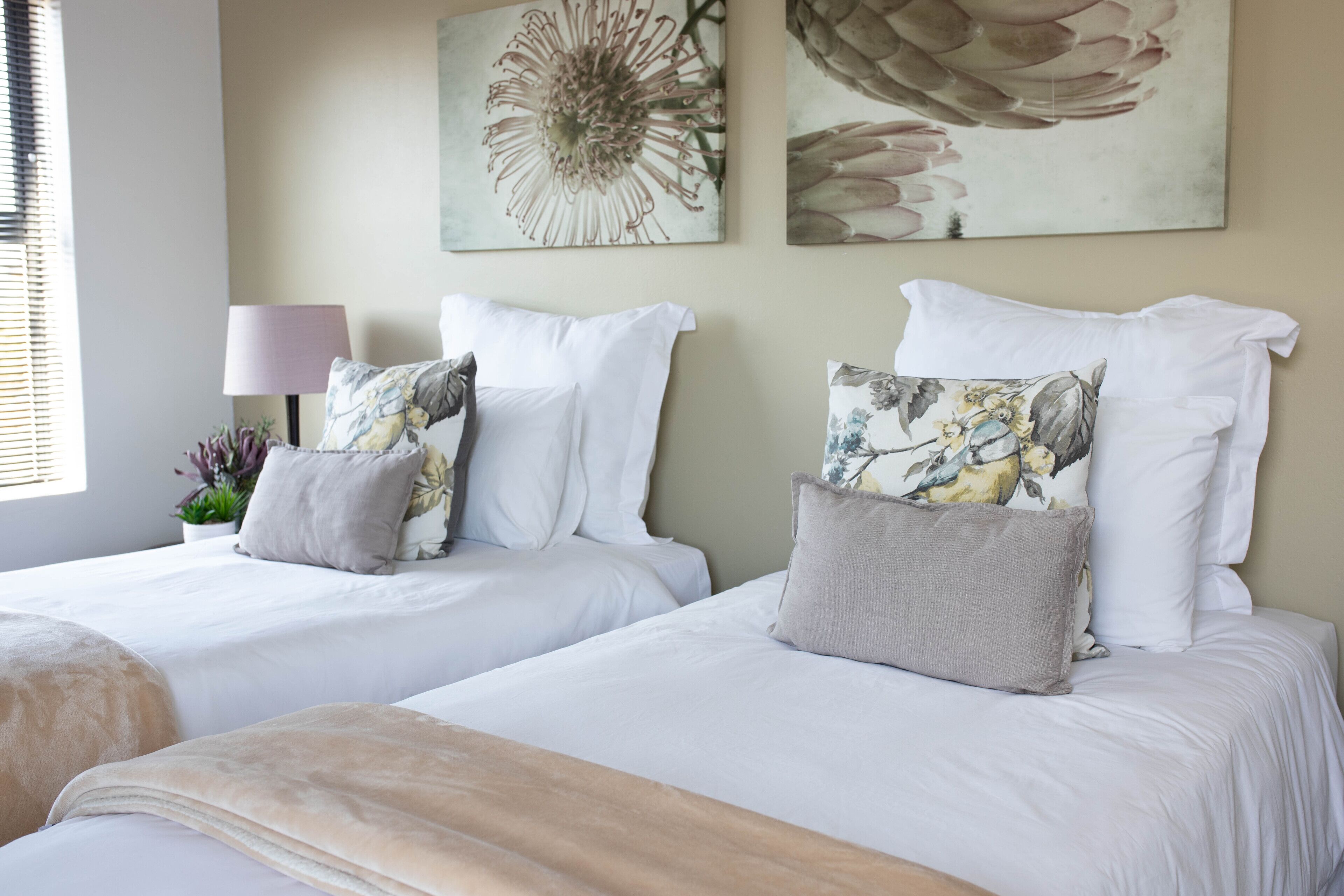 A hotel room with two beds, each with white bedding and decorative pillows. There is a lamp and a potted plant on the left side.