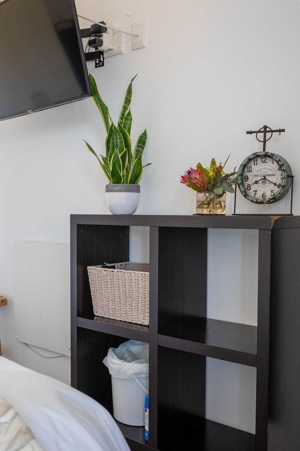 A room with a bed, a TV mounted on the wall, a black shelf with a woven basket and a plastic bag, and a wall clock.