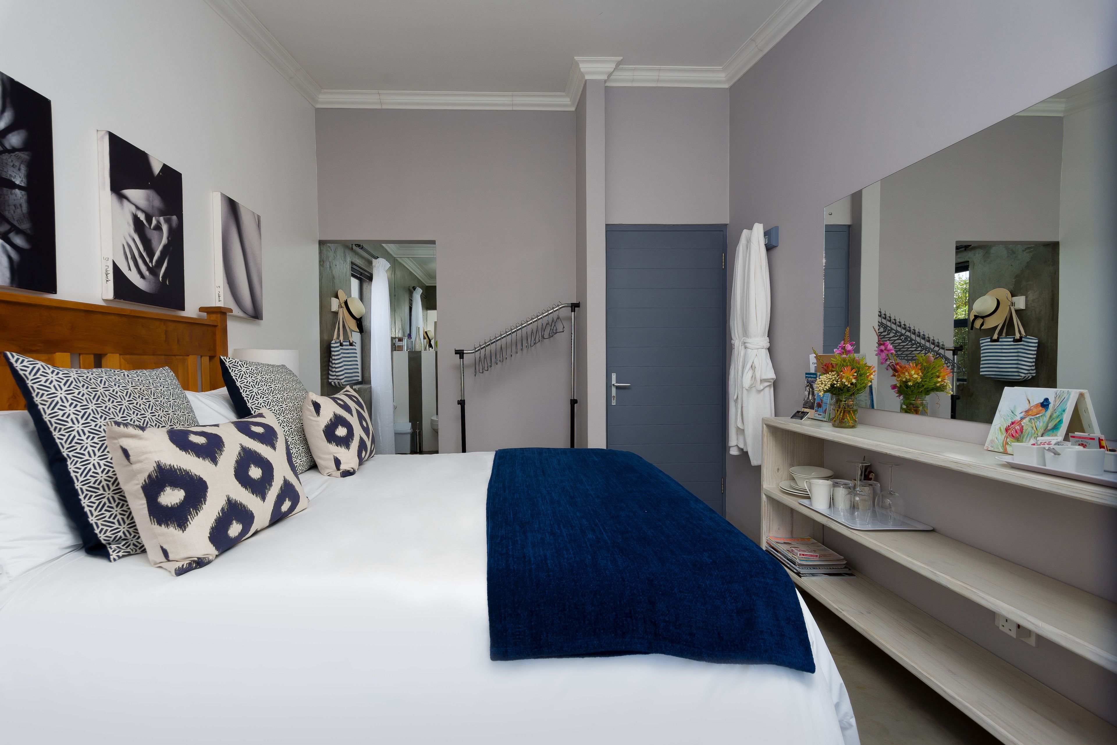 Un dormitorio con cama, almohadas, una manta azul, un espejo, un estante con artículos de aseo y una puerta que da a otra habitación.