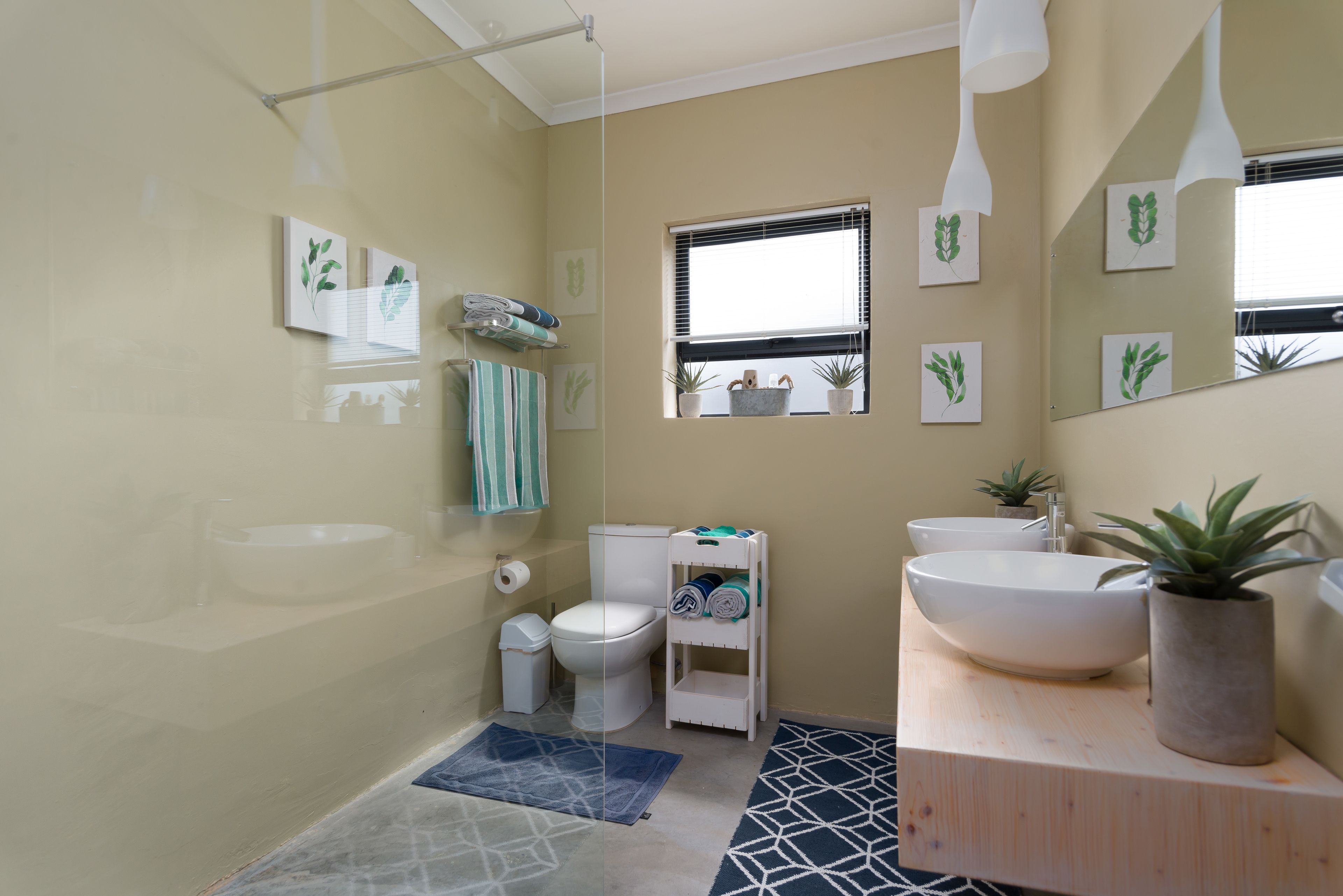 Une salle de bain avec un lavabo, des toilettes et un bidet blancs, une petite fenêtre avec des stores et un miroir agrémenté d’une plante.