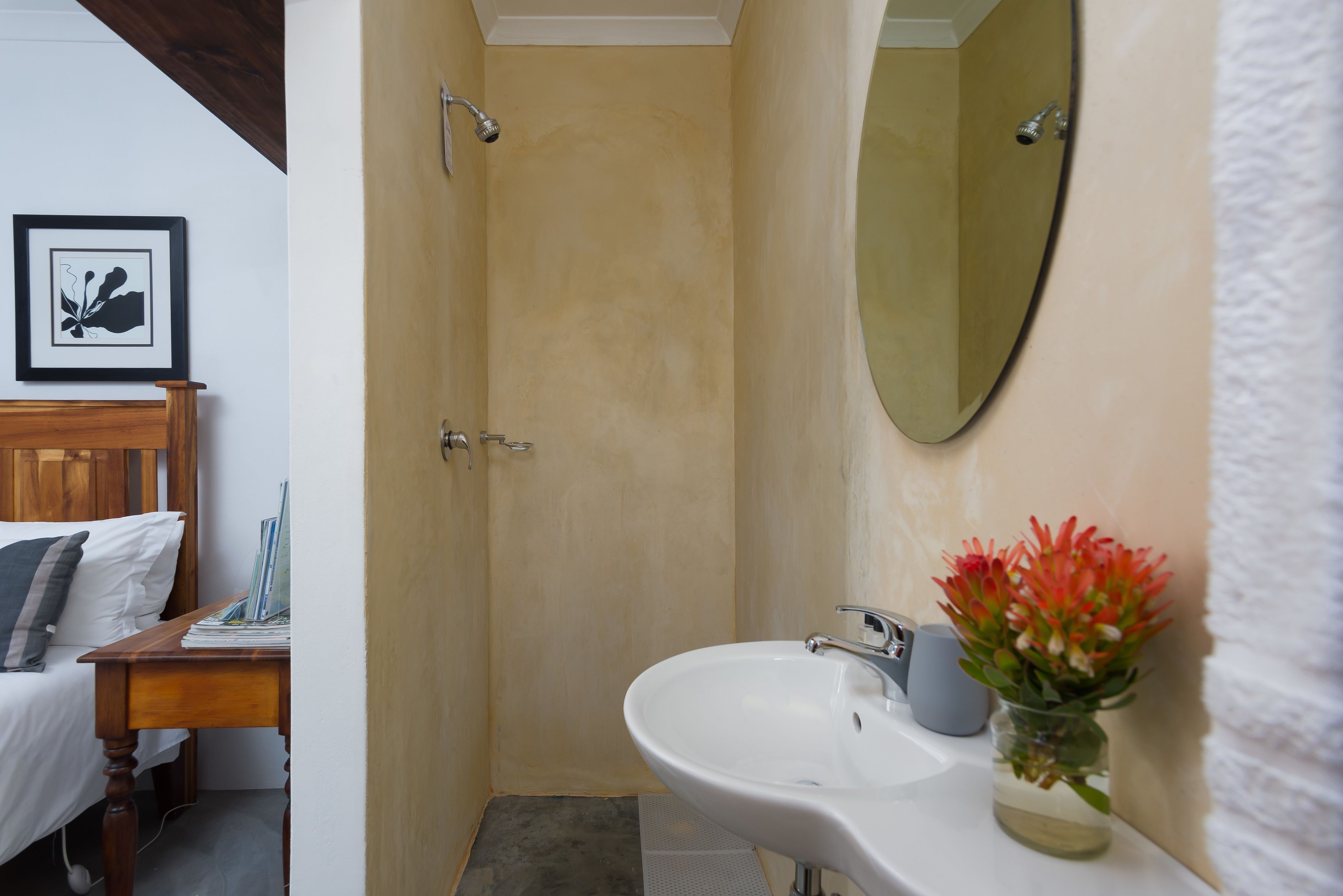 Un baño con ducha, lavabo y espejo. Hay una cama con almohadas y un cuadro en la pared.