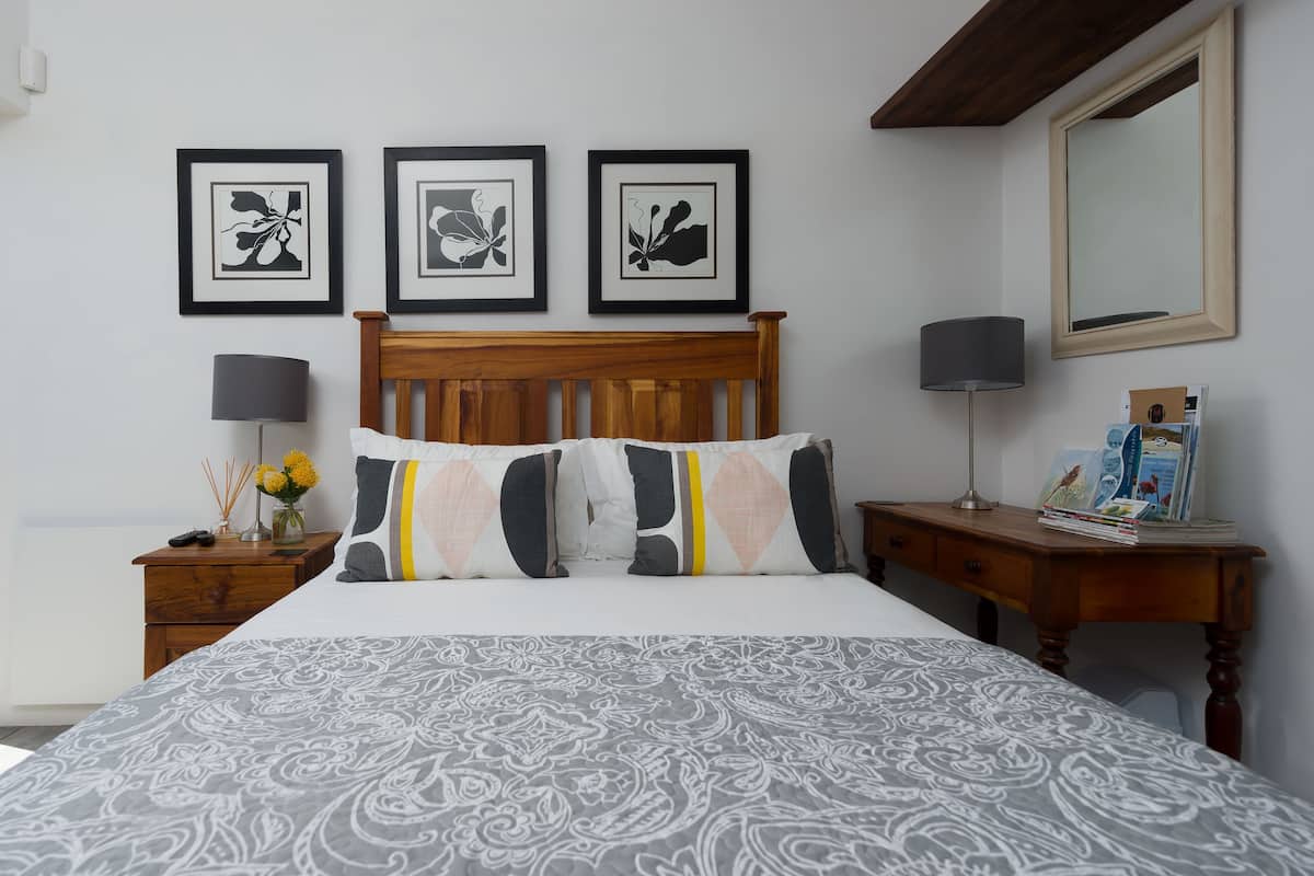 A bedroom with a wooden headboard, a patterned bedspread, a nightstand with a lamp, a dresser, and three framed floral artworks on the wall.