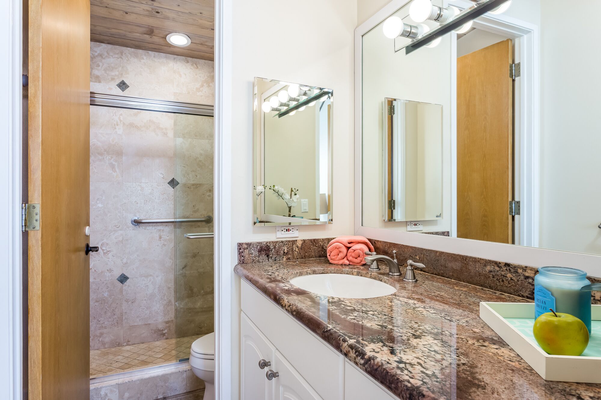 A bathroom with a glass shower enclosure, a sink with a granite countertop, and a mirror with lights above it.