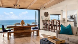 A spacious living area with a large window overlooking the sea, a wooden dining table, and a cozy seating area with a sofa and ottoman. - Seaside Luxury Duplex with Putting Green E950-0 (Encinitas)
