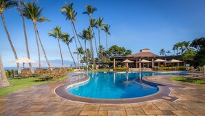 A swimming pool with lounge chairs and umbrellas, surrounded by palm trees and a building with a roofed area.