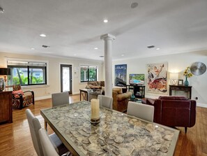 A spacious living area with a dining table, comfortable seating, and a large window offering a view of greenery. - Vidal Manor | Walk to Wilton Drive + Heated Pool (Wilton Manors)