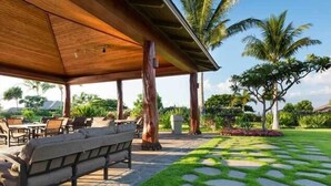 Un espacio de estar al aire libre cubierto con sofá y sillones, rodeado por un jardín con palmeras y un sendero de piedra.
