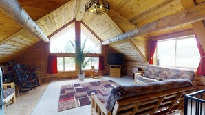 A cozy attic room with a wooden ceiling, a large window, a sofa, a TV, and a plant. - W-W: Canyon View Cabin, An Epic Family Retreat (Monticello)
