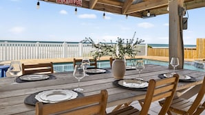 Un área de comedor al aire libre con mesas y sillas de madera, un letrero en lo alto y vista al océano.