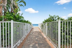 A paved pathway leading to a beach, flanked by white metal fences and lush greenery.