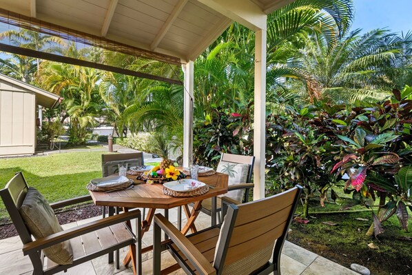 An outdoor patio with a wooden table set for dining, surrounded by chairs, and a view of a lush garden with various plants and a palm tree.