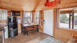 A wooden cabin interior with a kitchen area, a dining table, and a door leading outside.