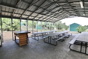 Un área de picnic techada con mesas y bancos de metal, una mesa de madera y piso de concreto.