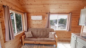 A cozy cabin interior with a wooden floor, a sofa with cushions, a window with curtains, and a kitchenette with cabinets.
