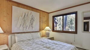 Een slaapkamer met een bed, nachtkastje, lamp, raam en een kunstwerk aan de muur.