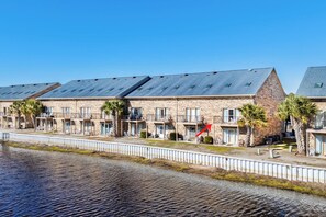 A row of stone buildings with balconies, a white fence, and a calm water body in the foreground.