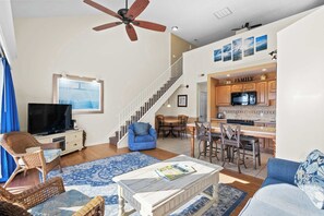 A spacious living area with a television, wicker furniture, a blue sofa, a coffee table, a ceiling fan, and a staircase leading to a kitchen area.