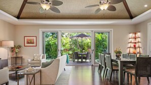 Ampla sala de estar com sofá, mesa de centro e mesa de jantar com cadeiras. Há ventiladores de teto, uma grande janela com vista para o verde, e uma prateleira decorativa com objetos vermelhos e laranja.