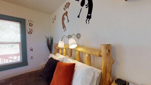 A bedroom with a wooden headboard, two bedside lamps, a bed with multiple pillows, a bedside table with a lamp, a small plant, and a wall with handprints and a stick figure drawing. - Hot Tub, Kokopelli Kabyn, Cute and Cozy! (Blanding)