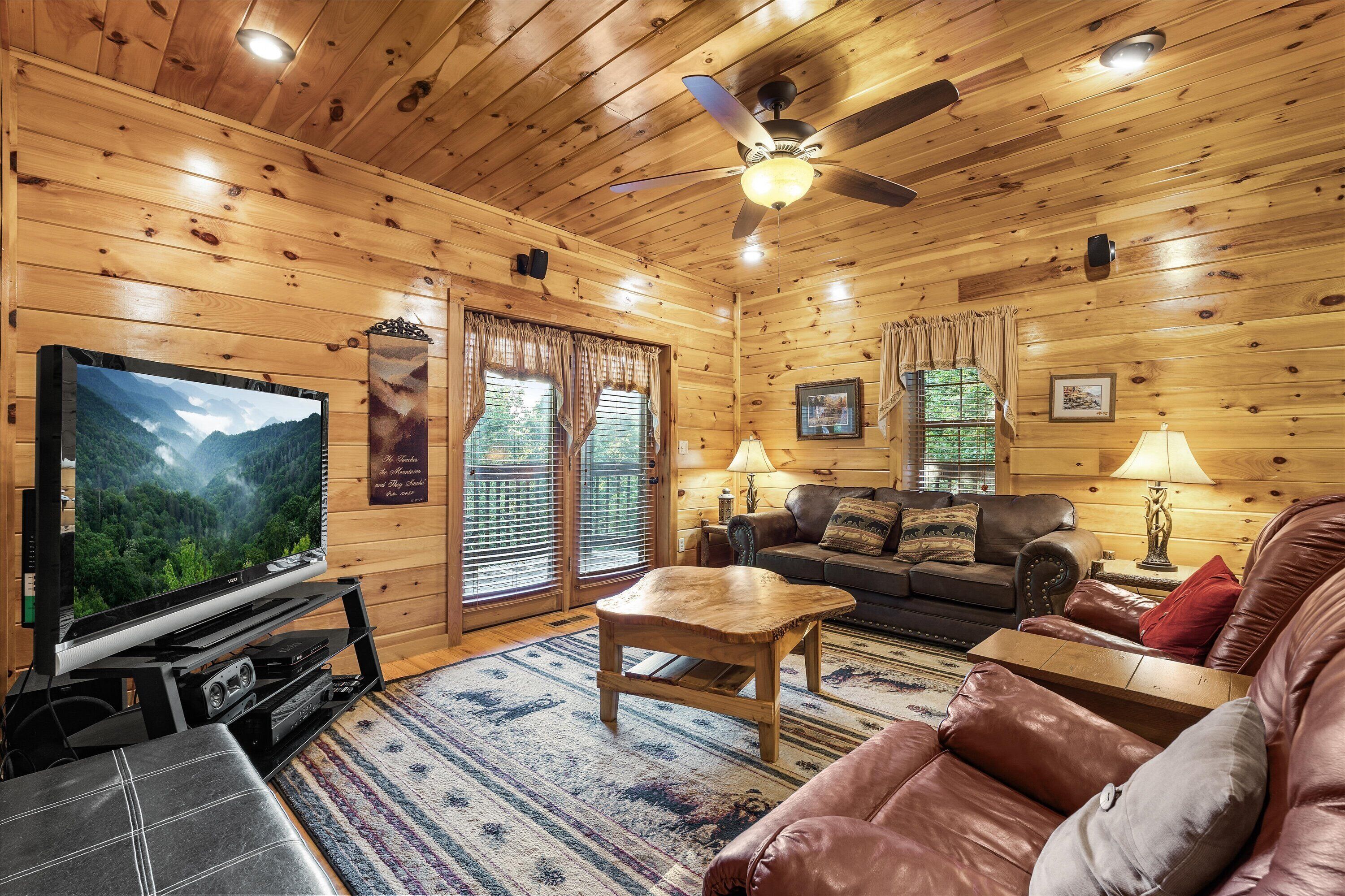 L’intérieur chaleureux d’un chalet avec un plafond en bois, un ventilateur de plafond, un téléviseur, un canapé en cuir, une table basse et une cheminée.