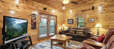 L’intérieur chaleureux d’un chalet avec un plafond en bois, un ventilateur de plafond, un téléviseur, un canapé en cuir, une table basse et une cheminée.