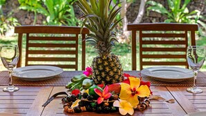 Una mesa de madera dispuesta para dos, con una piña como pieza central, rodeada de frutas y flores tropicales.