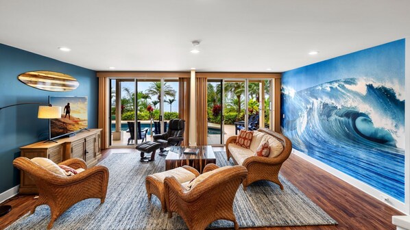 A spacious living area with wicker furniture, a large ocean wave mural, and a view of a pool and greenery.