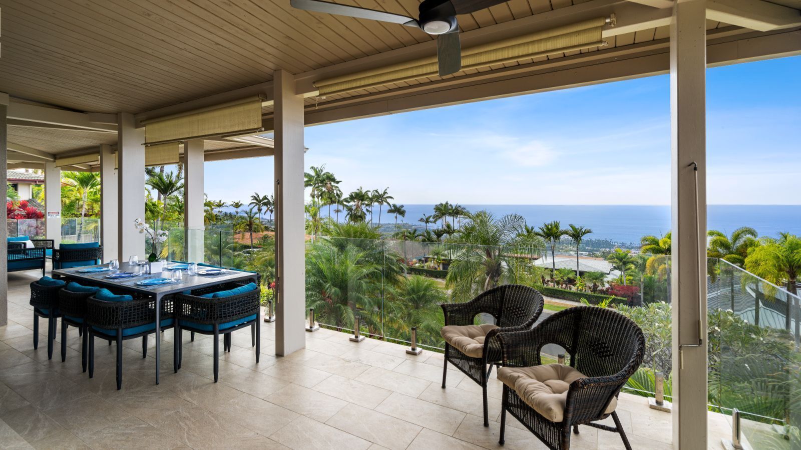 A covered outdoor dining area with a table and chairs, overlooking a scenic view of the ocean and palm trees.