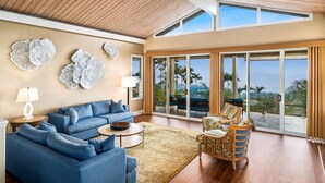 A spacious living room with a blue sofa set, a coffee table, and a wicker chair. Large windows offer a view of palm trees and the ocean.