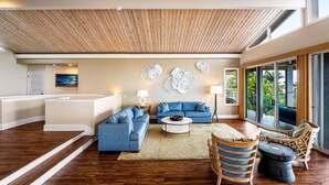 A modern living room with a blue sofa, a coffee table, and a wicker chair. There is a large window with a view of greenery outside.