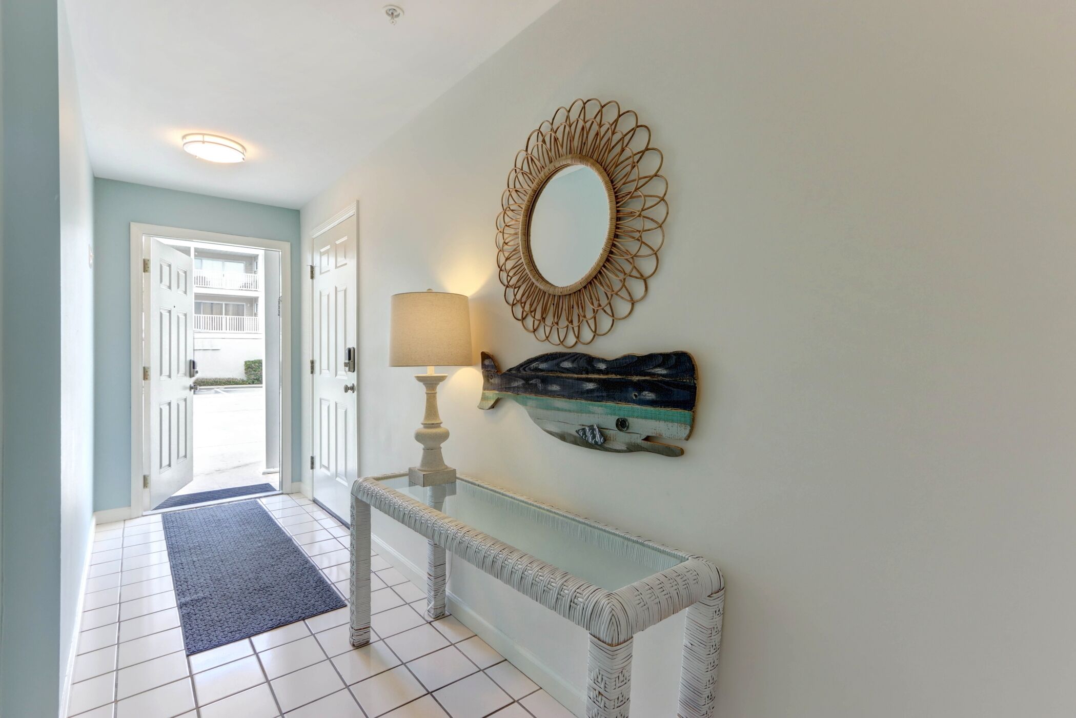 A hallway with a white tiled floor, a blue mat, a white console table with a lamp, and a decorative mirror on the wall.