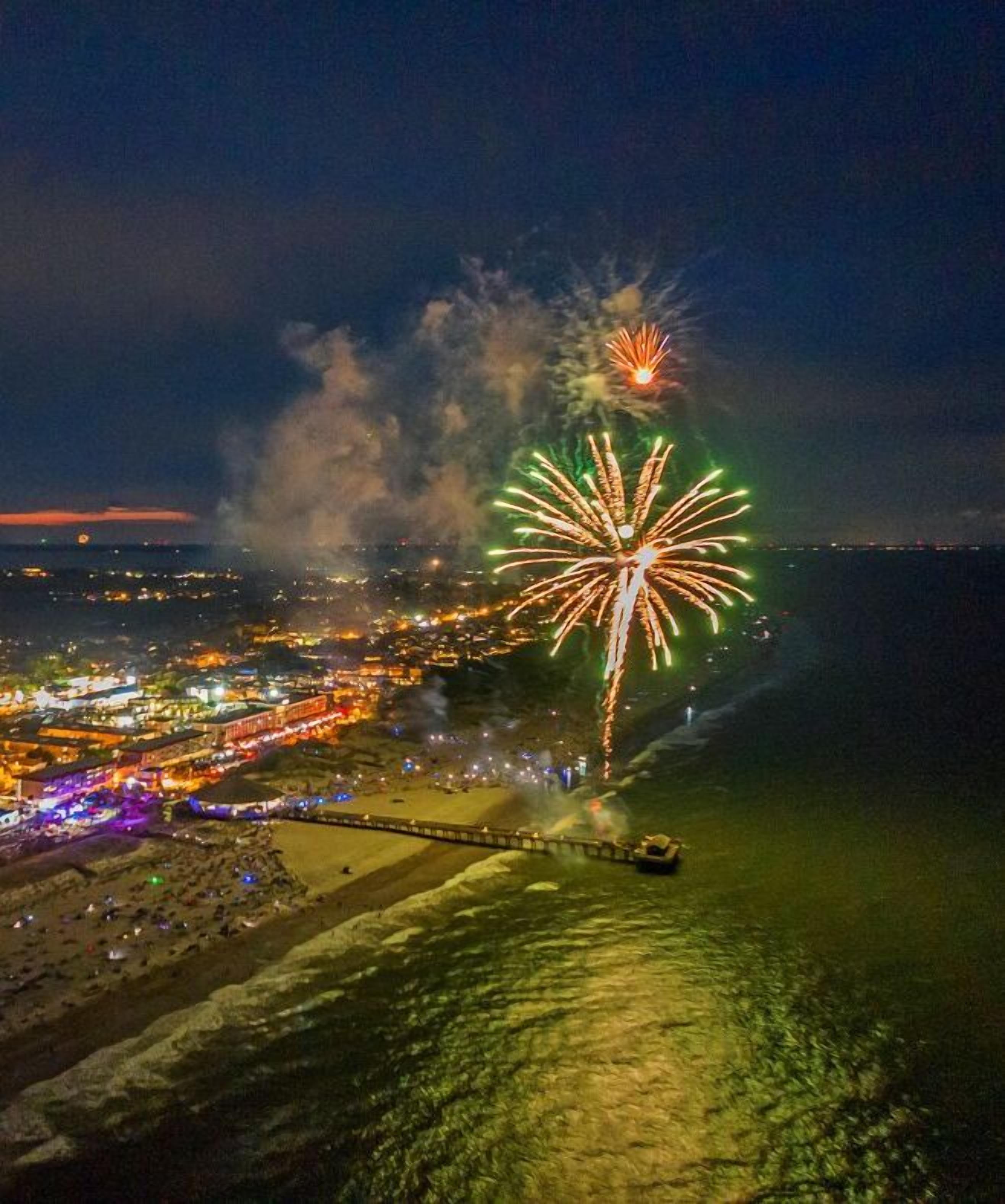 An aerial view of a coastal city at night with fireworks display over the water.