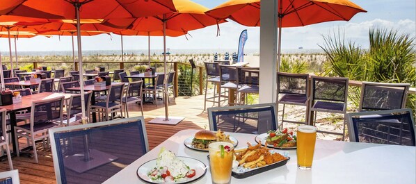An outdoor dining area with tables and chairs, umbrellas, and a view of the beach.