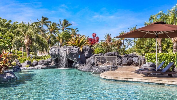 A clear pool with a waterfall feature, surrounded by tropical vegetation and lounge chairs.