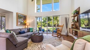 A spacious living room with a large sofa, a coffee table, and a rocking chair. There is a television on a wooden stand and a large window providing a view of the outdoors.