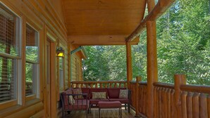 Eine hölzerne Veranda mit einer Bank und Kissen, umgeben von einem Holzaushandlauf, mit Blick auf eine bewaldete Landschaft.
