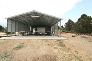 A covered outdoor area with picnic tables and a door, surrounded by trees and a gravel ground. - W-W: Long View "Safari" Cabin Overlooking Canyon! (Monticello)