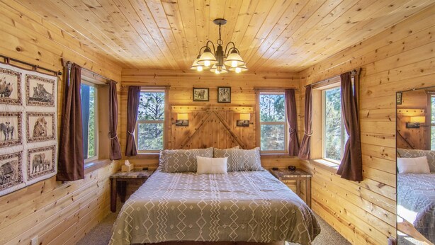 A wooden cabin bedroom with a large bed, a chandelier, framed pictures on the wall, and a window with curtains. - W-W: Long View "Safari" Cabin Overlooking Canyon! (Monticello)