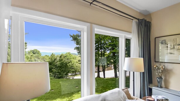 A room with a large window offering a view of a grassy area and trees, a beige sofa with cushions, and a lamp on a side table. - Bright mountain condo w/ fireplace near Sugarbush (Warren)