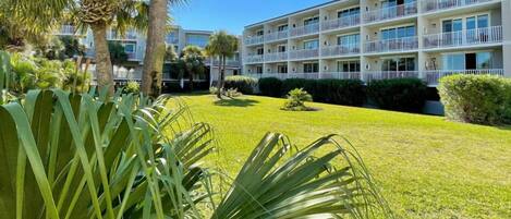 A multi-story hotel with balconies, a well-maintained lawn, and tropical plants.
