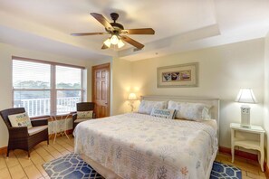 A bedroom with a bed, a ceiling fan, a window with blinds, a chair, a nightstand, and a lamp.