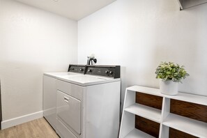 Une buanderie équipée d’un lave-linge et d’un sèche-linge à chargement frontal, d’une étagère en bois et d’une plante en pot.