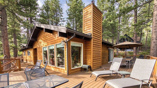 A wooden cabin with a deck, furnished with metal chairs and tables, surrounded by tall pine trees.