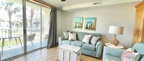 Sala de estar con ventilador de techo, sofá, mesa de centro y lámpara. Hay dos cuadros en la pared y una ventana con vistas a palmeras.